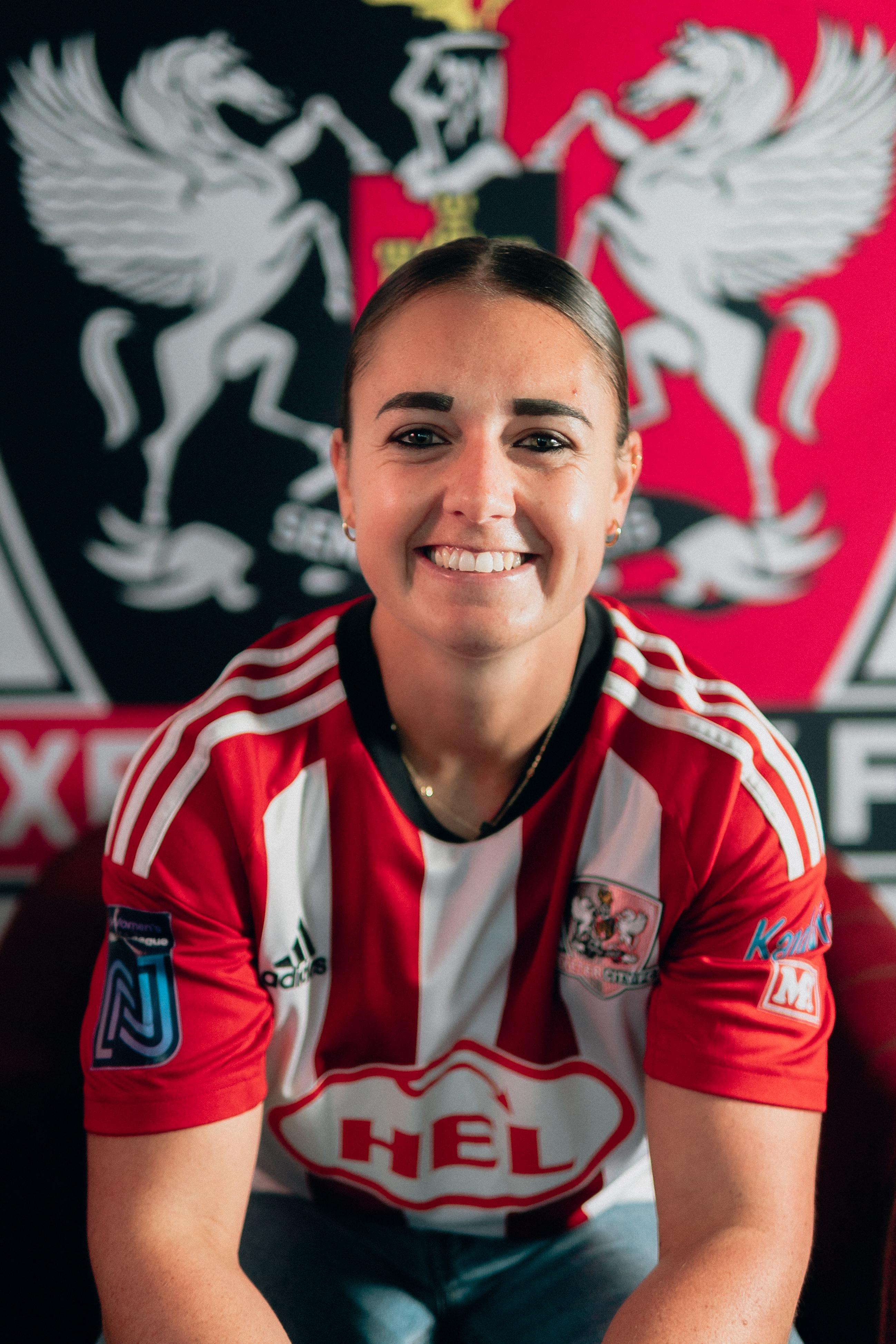 Image of Leah Burridge sat down wearing a red and white Exeter City home shirt in front of the Exeter City badge