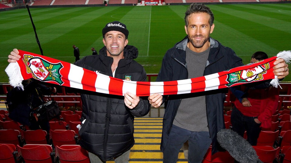 Rob McElhenney and Ryan Reynolds holding a Wrexham scarf aloft inside the Racecourse Ground