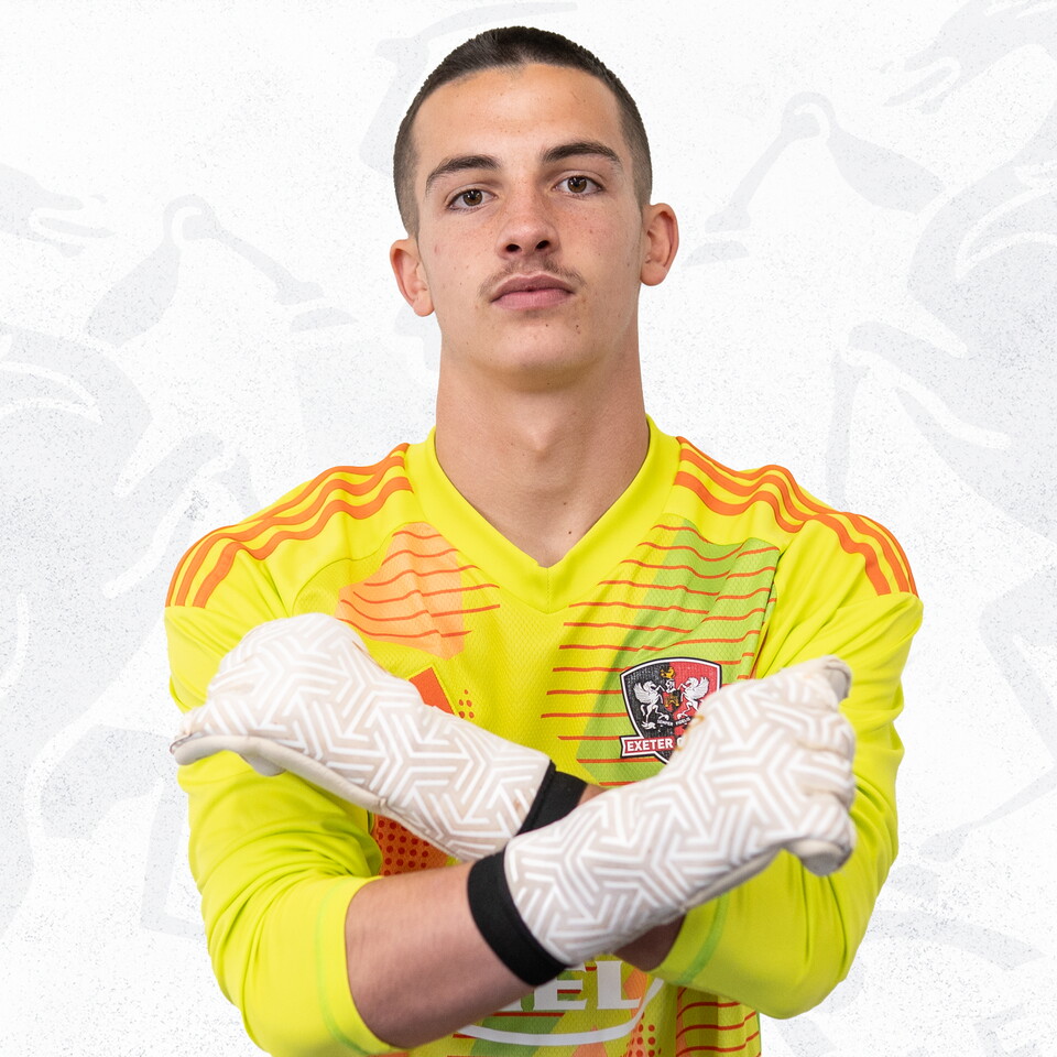 Dylan Shanahan profile picture. He's wearing a bright yellow Exeter City goalkeeper top, and has his arms crossed in an 'X' position, with his goalkeeper gloves on. He's got short dark hair and a light moustache.