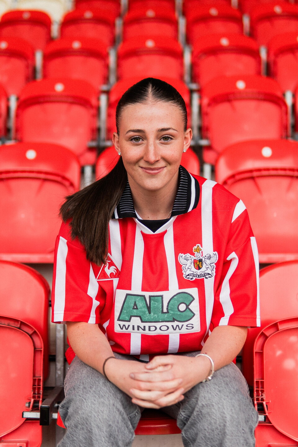 A photo of Georgia Vaccaro sat in the red seats of the Adam Stansfield stand wearing a retro Exeter City shirt